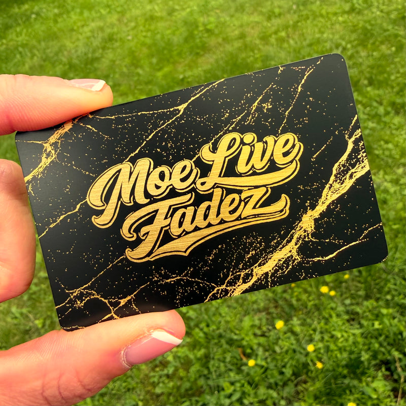 Black brass tap NFC card with gold marble pattern and 'Moe Live Fadez' text held by a hand against a grassy background